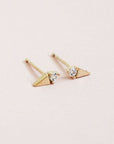 In front of a soft pink background is a pair stud earrings. On one side of each stud is a gold triangle and on the other side is a white diamond. The diamond is on the flat side of the triangle. 