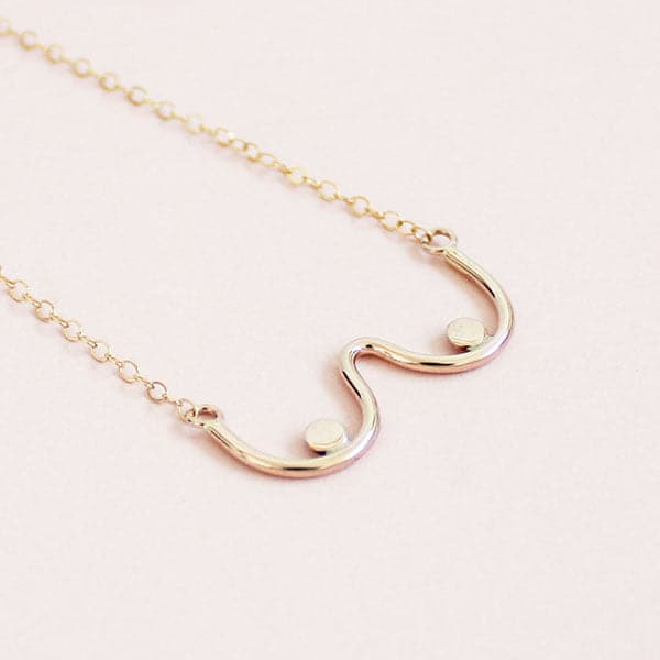 A gold chain necklace with a dainty pendant shaped like the outline of boobs.