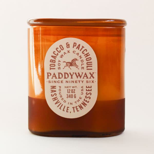 Oval cylinder shaped glass candle with bright tones of burnt orange. The label is an oval shape and reads 'Tobacco & Patchouli' with a galloping horse image within. The middle reads 'PaddyWax Since Ninety Six: Nashville, Tennessee'. 