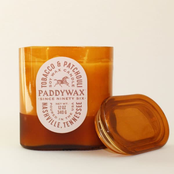 Oval cylinder shaped glass candle with bright tones of burnt orange. The label is an oval shape and reads &#39;Tobacco &amp; Patchouli&#39; with a galloping horse image within. The middle reads &#39;PaddyWax Since Ninety Six: Nashville, Tennessee&#39;. Alongside its lid sits propped up against the candle. 