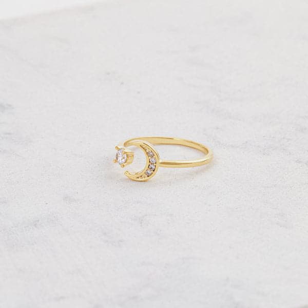 A gold ring with an open front that features a cubic zirconia on one end and a crescent moon on the other, photographed worn on a models hand.