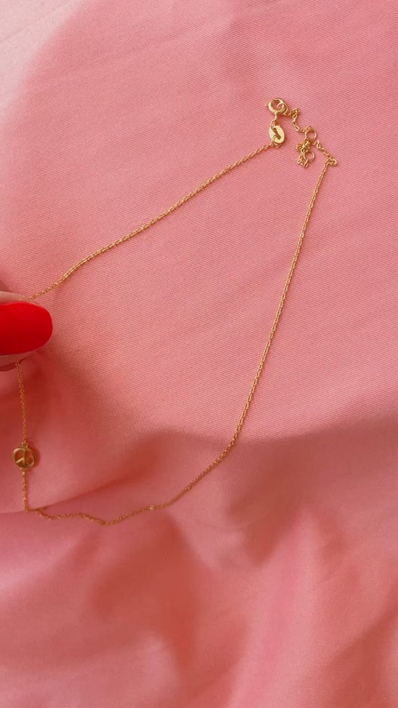 A video of a hand picking up and setting down a gold necklace with a sample pace sign pendant at center.  The necklace sits on top of a soft pink textured fabric.