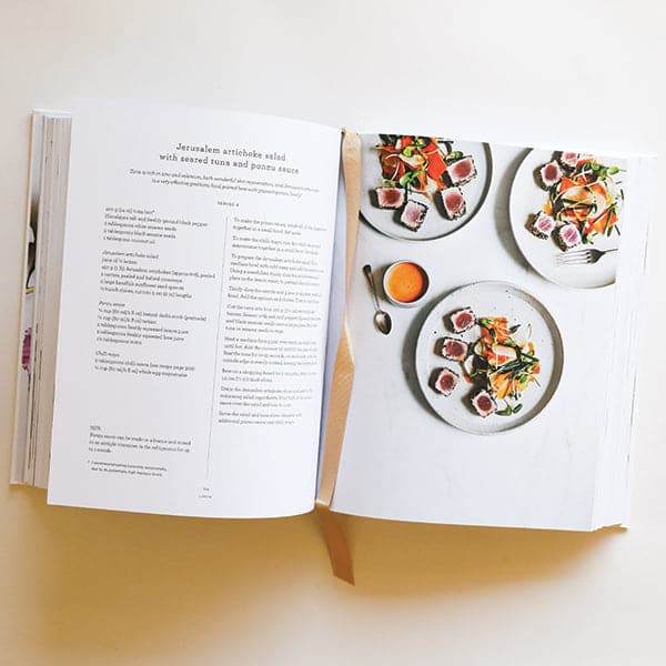 Two open pages of the cookbook. To the left is a detailed recipe in black text and to the left is three plates filled with vibrant artichoke salad with fresh seared tuna The three plates sit against a marbled table top and alongside tangerine colored dressing. 