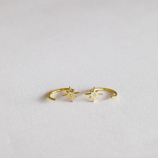 In front of a white background is a pair of tiny, half hoop gold earrings. On one end of both earrings is a small, gold star with a small clear, crystal in the middle. 