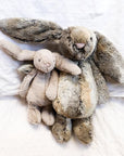 Two, on small and one large, stuffed animal in the shape of rabbits laying on a bed. The small rabbit is beige colored, and the large rabbit is mottled grey and gold.