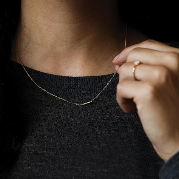 Photo of a woman with dark hair and a dark grey knit top wearing a thin gold necklace with a gold bar and CZ accents. Her hand is up by her neck and she is holding the chain between her pointer finger and thumb. She is wearing a small gold and opal ring on her ring finger. 