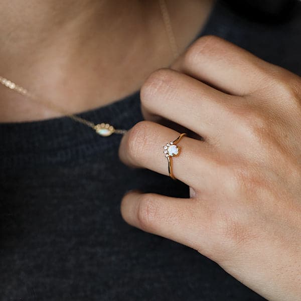 This is a close up picture of a woman’s chest. She is wearing a charcoal gray shirt. Her hand is pulling on a gold chain necklace around her neck. On her ring finer is a gold ring. On the front is a round white opal with five white stones on the top of the opal. 