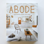 A book lays on a table titled "Adobe - thoughtful living with less." Cover shows inside a bright country - boho style home.