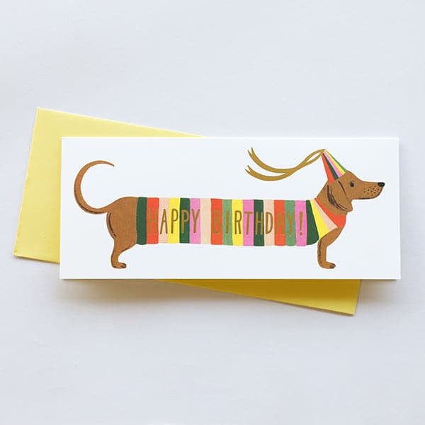 A long thin birthday card with a wiener dog illustration on the front with a multicolored stripe sweater on with gold lettering across it that reads, "Happy Birthday". The dog is also wearing a multicolored birthday hat with two ribbons hanging from it. The card comes with a yellow envelope photographed here.