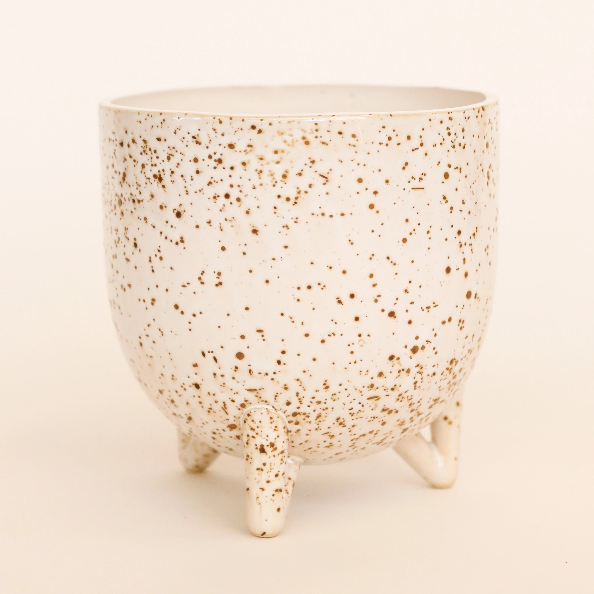 In front of a white background is a round footed pot. The pot is a cream color with dark brown specks. Seen from this view is three of the four feet at the bottom of the pot.