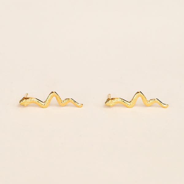 In front of a tan background is a pair of gold snake stud earrings. The post for the studs sticks out of the snakes head.
