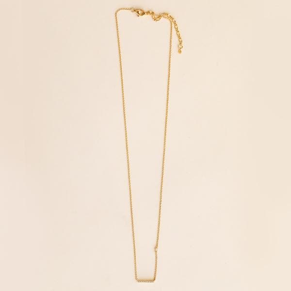 Photo of a gold necklace on a light pink background. Necklace is a gold chain with a gold bar with CZ accents.