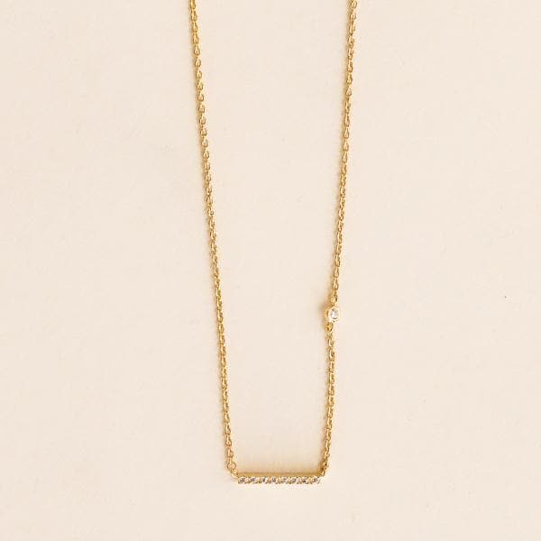 Close up photo of a gold necklace on a light pink background. Necklace is a gold chain with a gold bar with CZ accents. 
