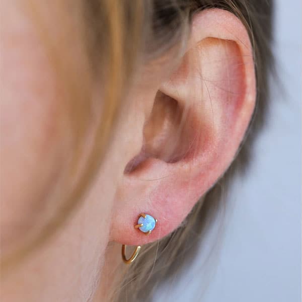 This is a close up picture of a woman’s ear. She is wearing a pair of gold huggies with a round green opal at the top. 