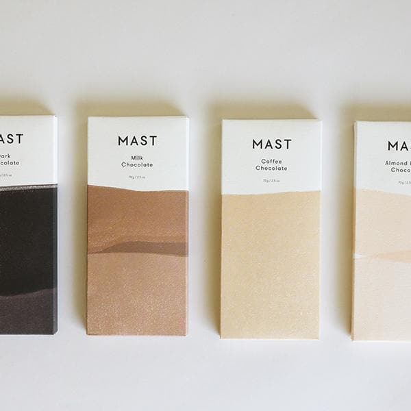 A rectangular bar of chocolate that read, "Mast Milk Chocolate" at the top in black letters along with a two toned wrapper that is brown and white alongside other flavors of chocolate by the same brand that feature different color ways.