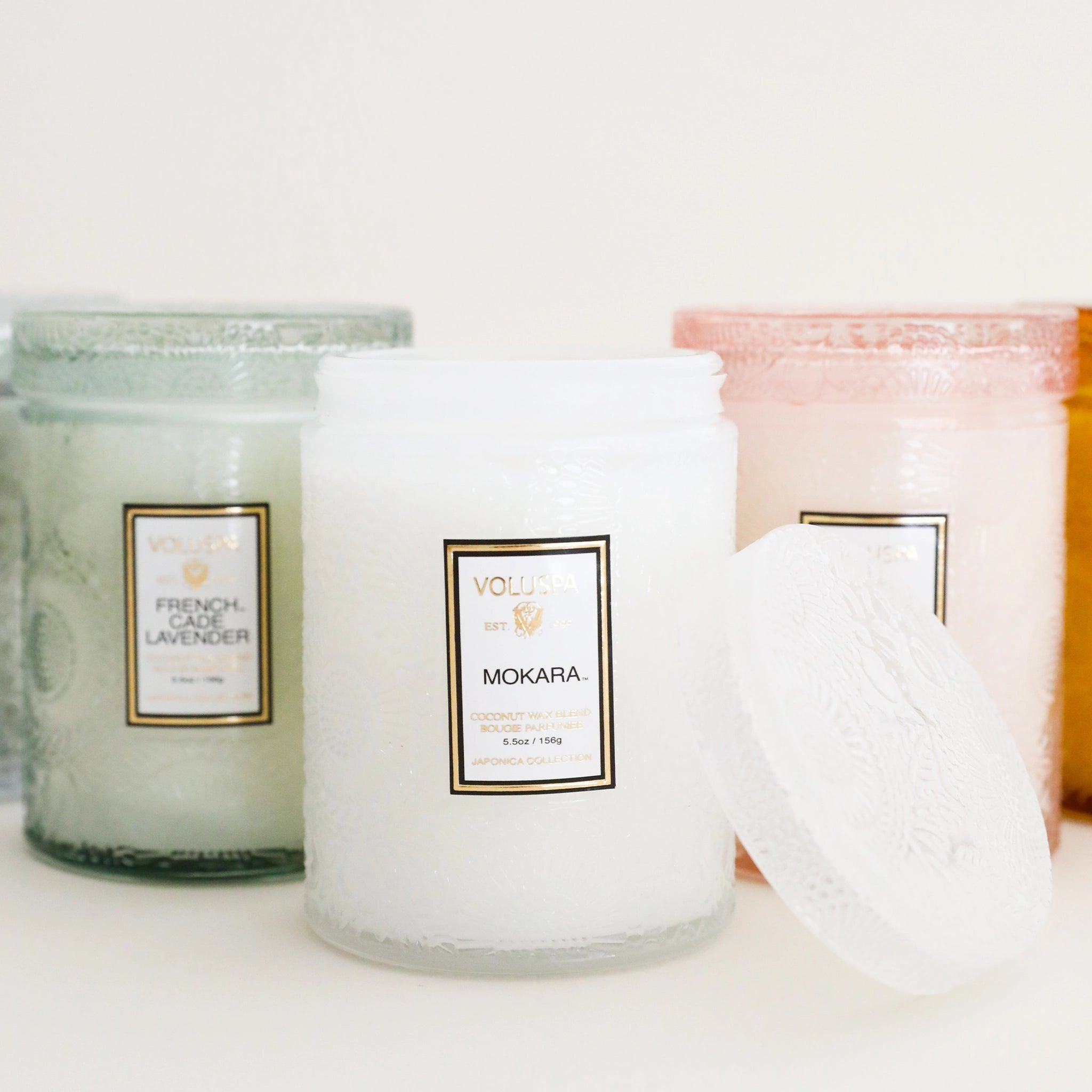 A decorative glass jar candle in a white color with a rectangular label on the front that reads, "Voluspa Mokara" alongside other scents of the same brand candle that feature different color ways.