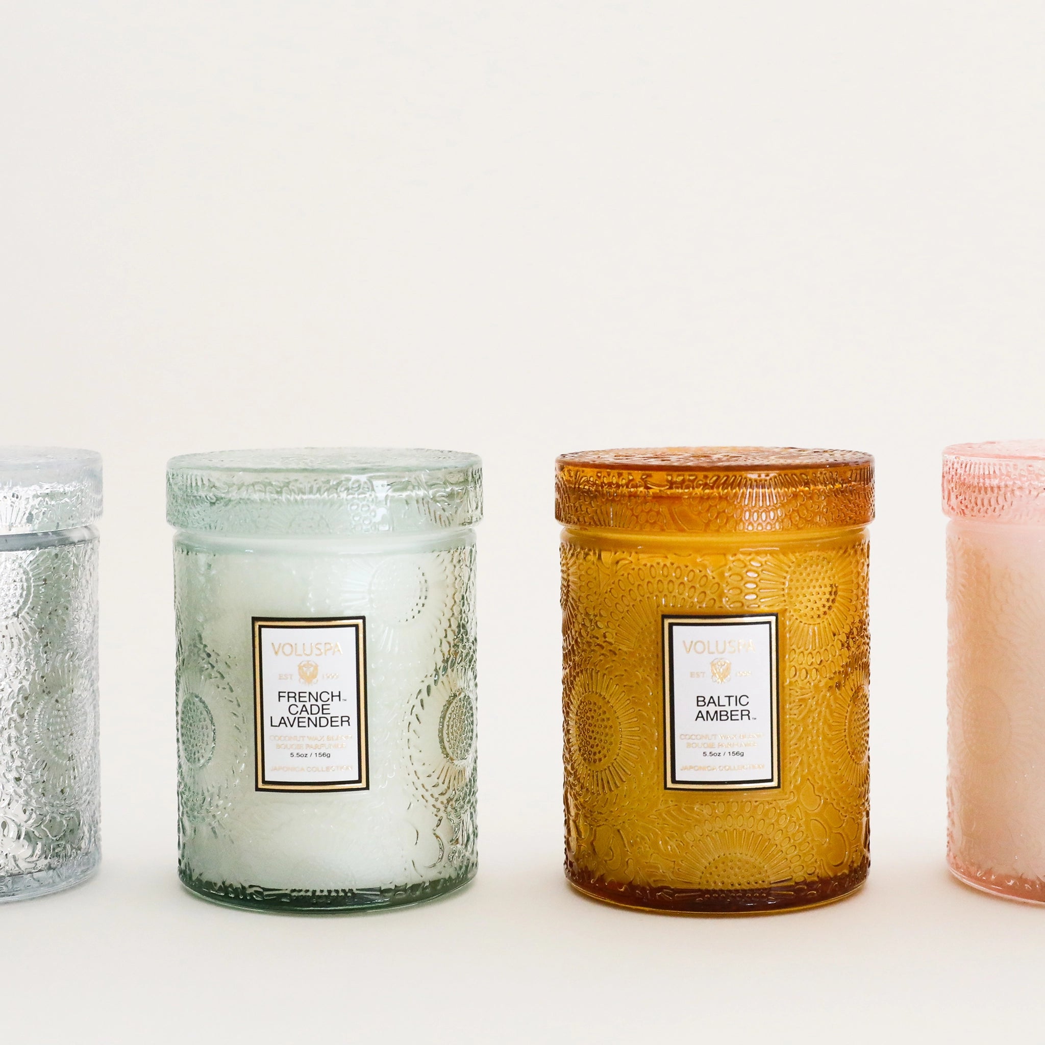 A light green decorative glass jar candle with a white rectangular label that reads, "French Cade Lavender" next to other scents by this brand that our store sells, like Baltic Amber which comes in a burnt orange glass jar.