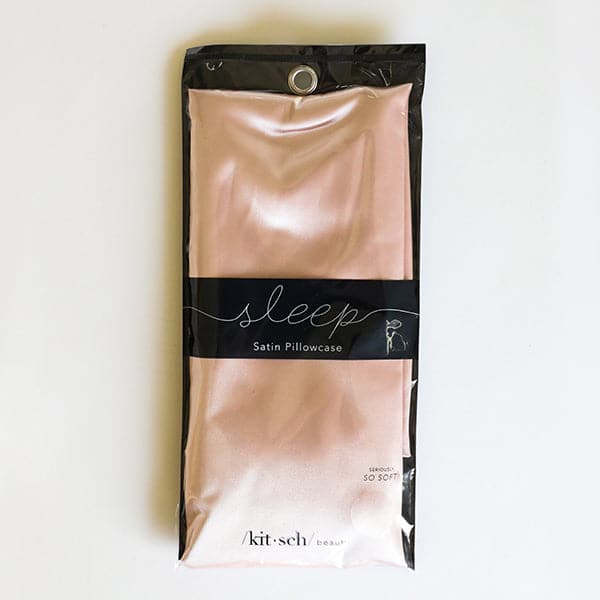 In front of a white background is a rectangular black package with a clear top. Inside the package is a folded satin pillowcase. The pillowcase is blush pink. There is a black horizontal band around the middle with white text that reads ‘sleep satin pillowcase.’ 