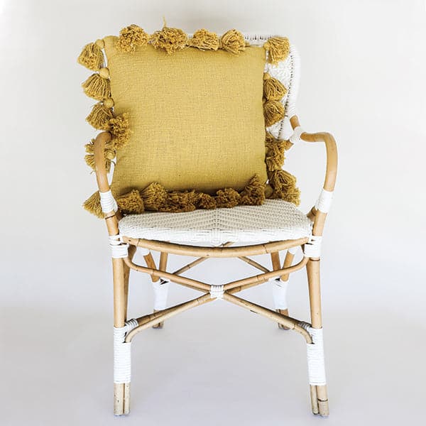 Against a white background is a white and brown wicker chair. On the chair is a mustard colored square pillow. The pillow is lined with small balls with tassels. 