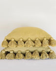Against a white background is a stack of two yellow, square pillows. The edge of both pillows are lined with small yellow balls with tassels. 