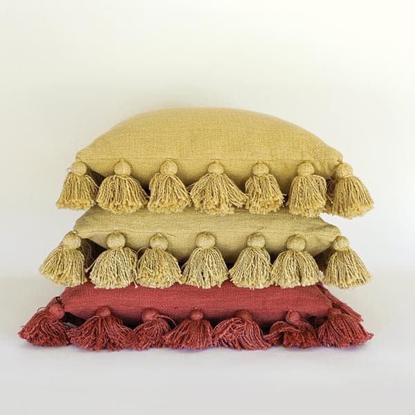 Against a white background is a stack of three square pillows. The edge of all of the pillows are lined with small  balls with tassels. The top two pillows are mustard and the bottom pillow is rust.