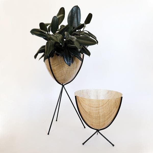In front of a white background is a wood colored bullet planter. The planter has a wide top and narrows at the bottom. The planter is held up by a black metal stand. The stand has three black metal legs. Inside the pot is a deep green plant. To the right is another wood colored bullet planter that is held up by a black metal stand. This one is about half the size of the one on the left. 