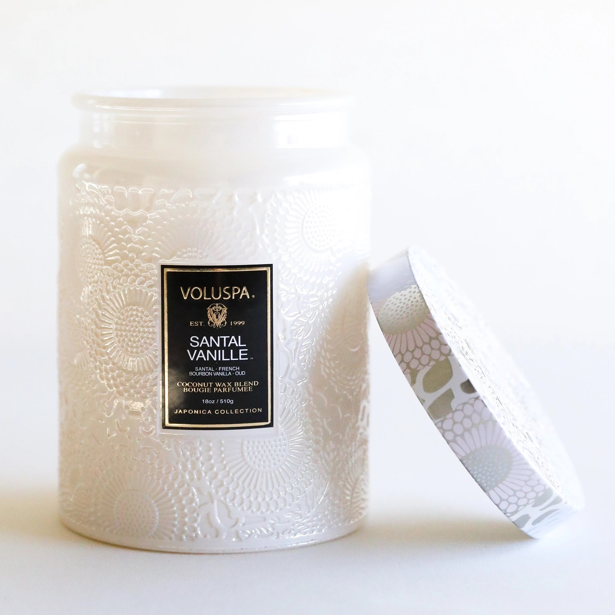 In front of a white background is a large light pink glass jar. The glass jar has a floral pattern all over it. In the middle is a black rectangular sticker with gold text at the top that reads ‘voluspa.’ Below is white text that reads ‘santal vanille.’ Leaning against the right side of the jar is a round tin lid. The lid has a light pink, silver and lavender flower pattern on it.