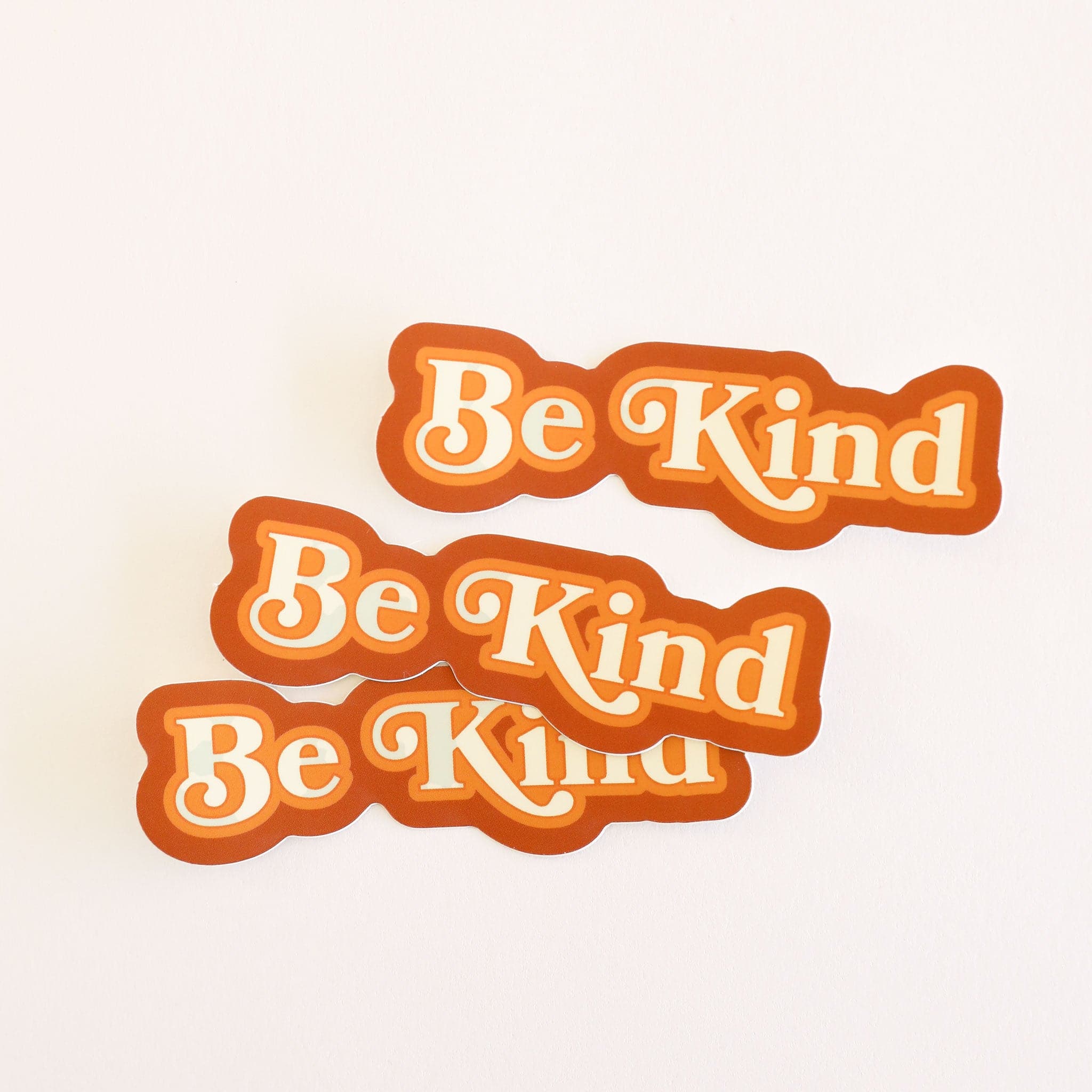 Three stickers reading &#39;Be Kind&#39; in white lettering, outlined in tangerine orange and rusty red.