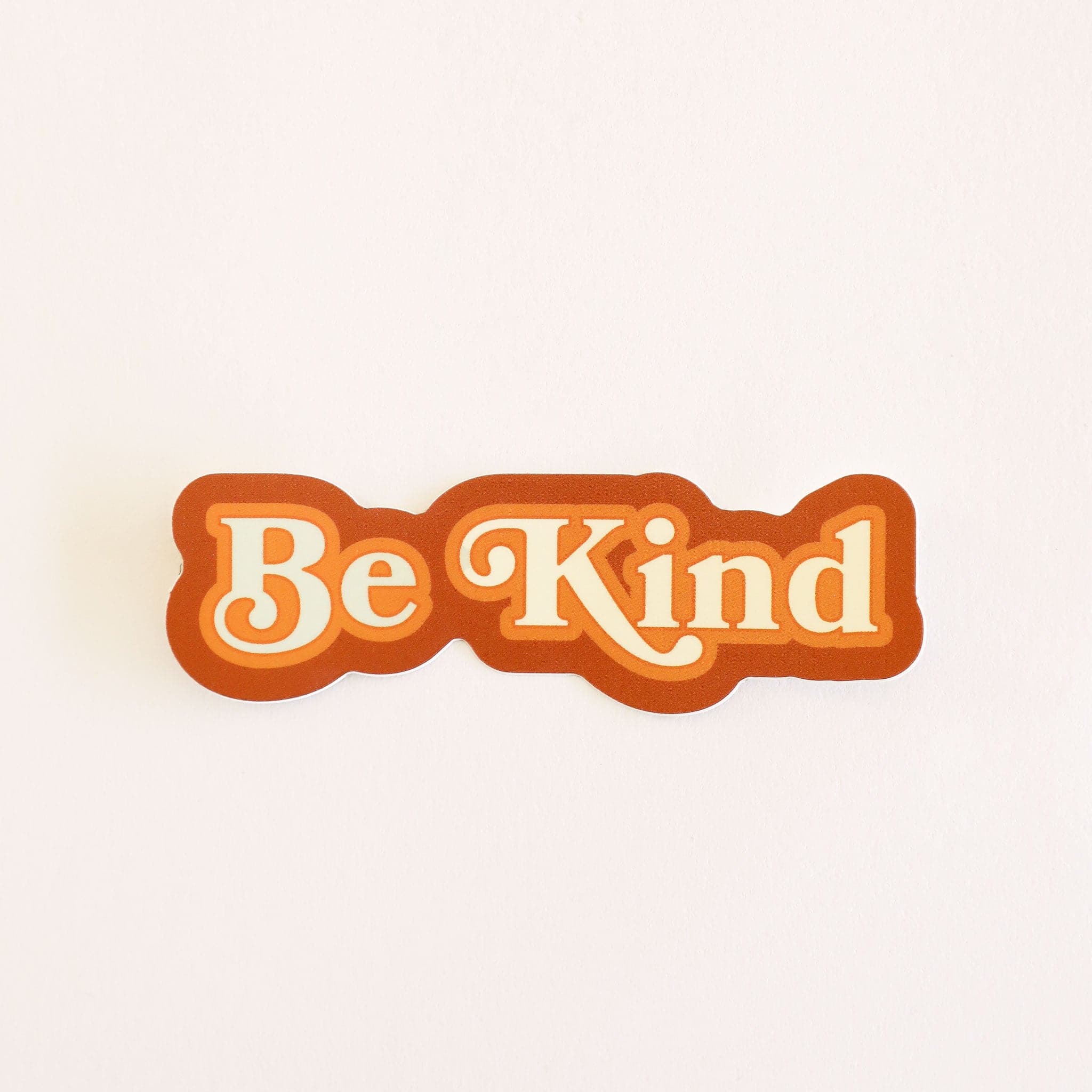 Sticker reading &#39;Be Kind&#39; in white lettering, outlined in tangerine orange and rusty red.