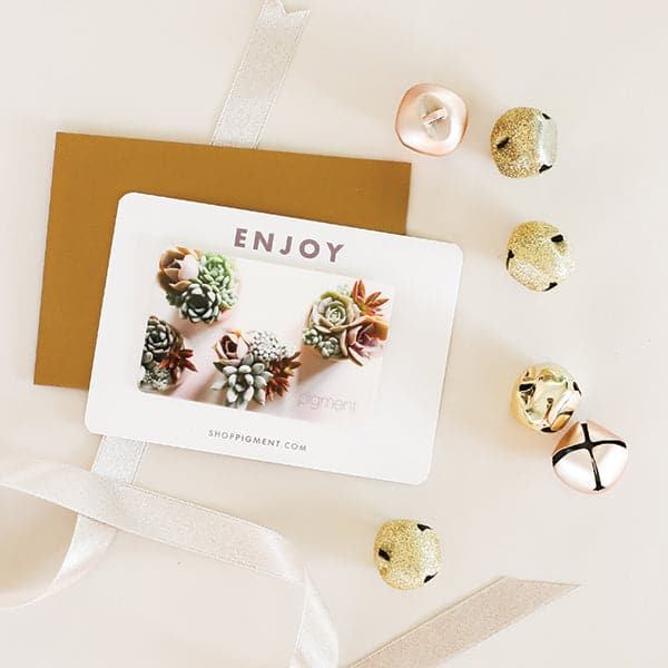 Birds eye view of a white background. Sitting on top of a gold envelope is a white gift card. At the top of the gift card is light purple text that reads ‘enjoy.’ Below the text is a birds eye view picture of four succulent arrangements. To the right of the gift card is six jingle bells. Three are glittery gold and the other three are rose gold.