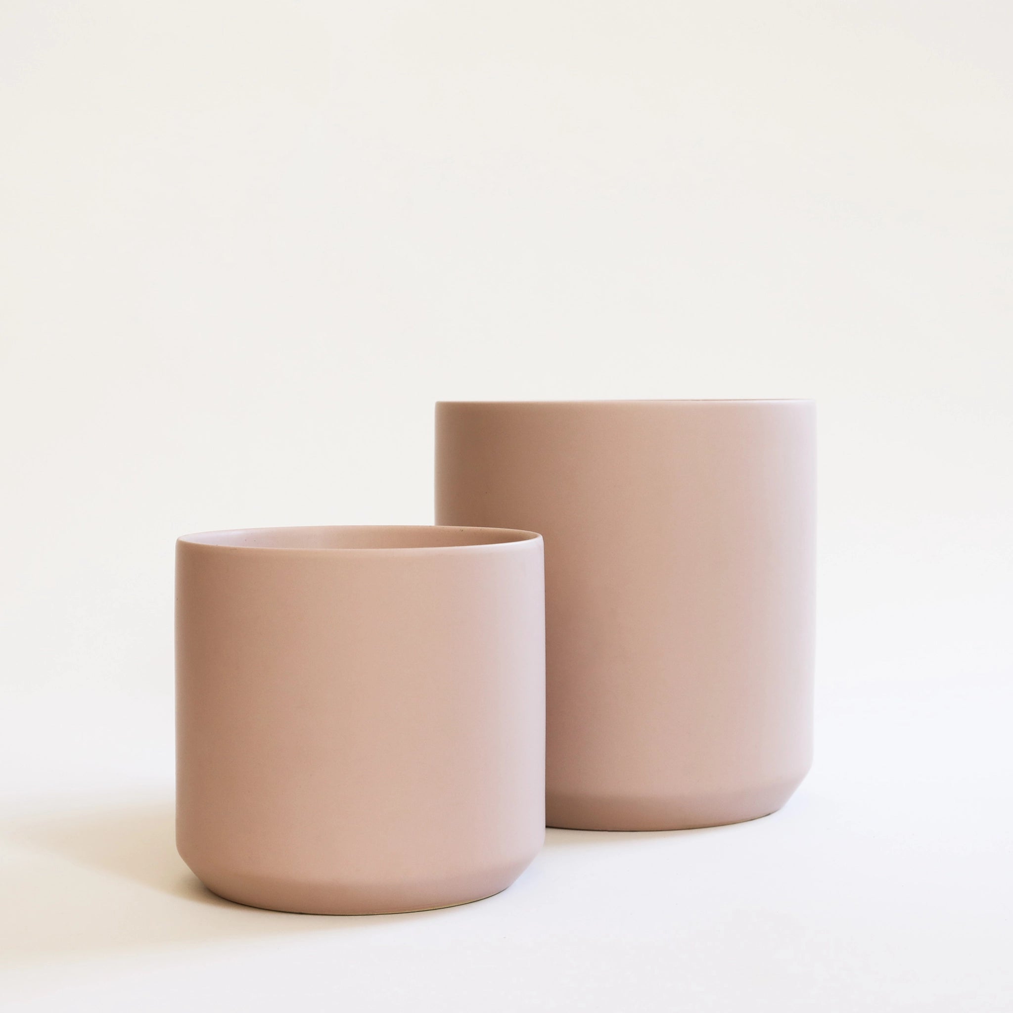 On a cream background is two different sized light pink ceramic planters. 