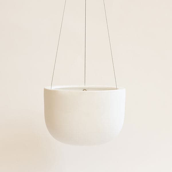 Hanging in front of a white background is a white hanging pot. The pot is wide and round and slightly tapers at the bottom. The pot is being held by three silver wires.