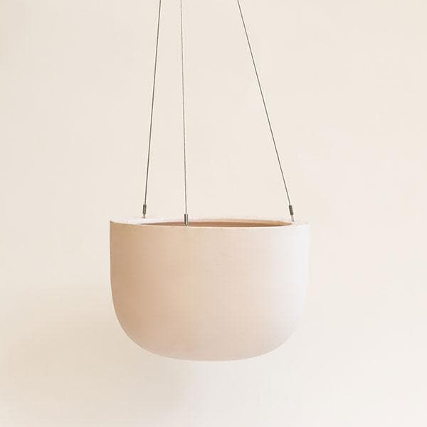 Hanging in front of a white background is a light pink hanging pot. The pot is wide and round and slightly tapers at the bottom. The pot is being held by three silver wires.