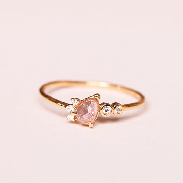Against a soft pink background is a slim, gold ring. On the front of the ring is a light pink crystal tear drop. Lining the left side of the tear drop are three white, round crystals. To the right of the tear drop is two white crystals with a gold border.