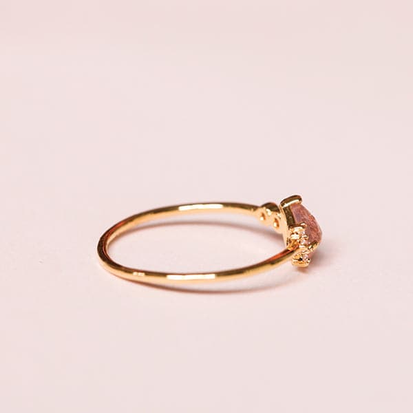 Against a soft pink background is the side view of a gold ring. The band of the ring is thin. Seen from this view is the side view of a soft pink tear drop crystal.