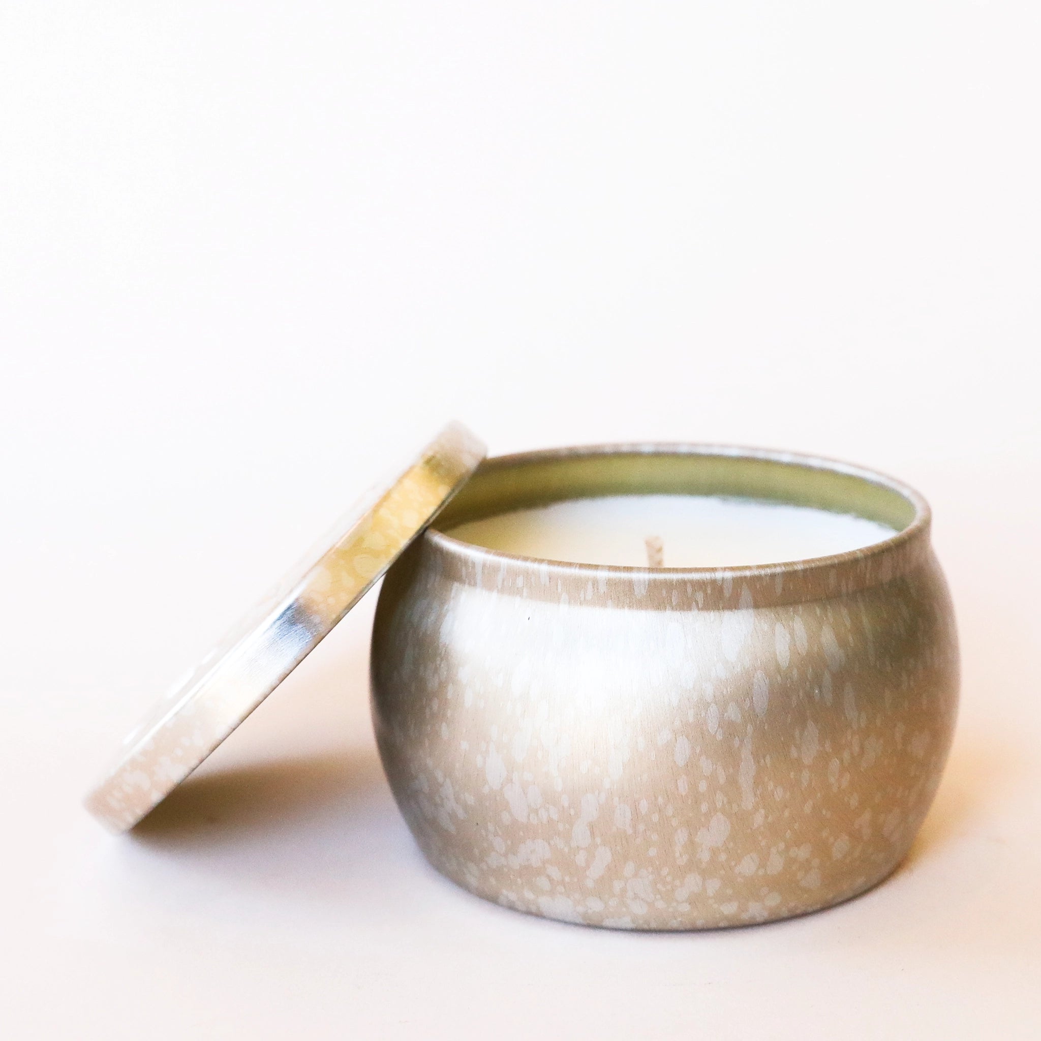 On a cream background is a light silver and gold mini tin candle with white wax and a single wick. Also included is the matching lid.