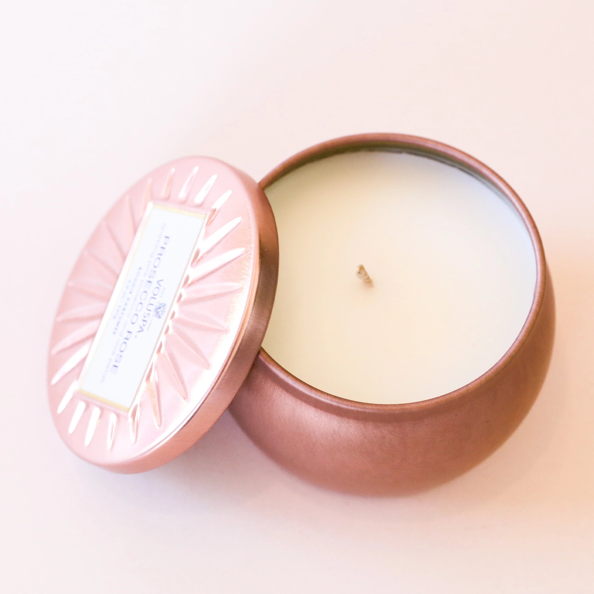 Birds eye view of a round rose gold tin candle. Inside is a white candle with a white wick. There is a matching lid leaning agains the left side. On the lid is a white rectangle with black text inside.