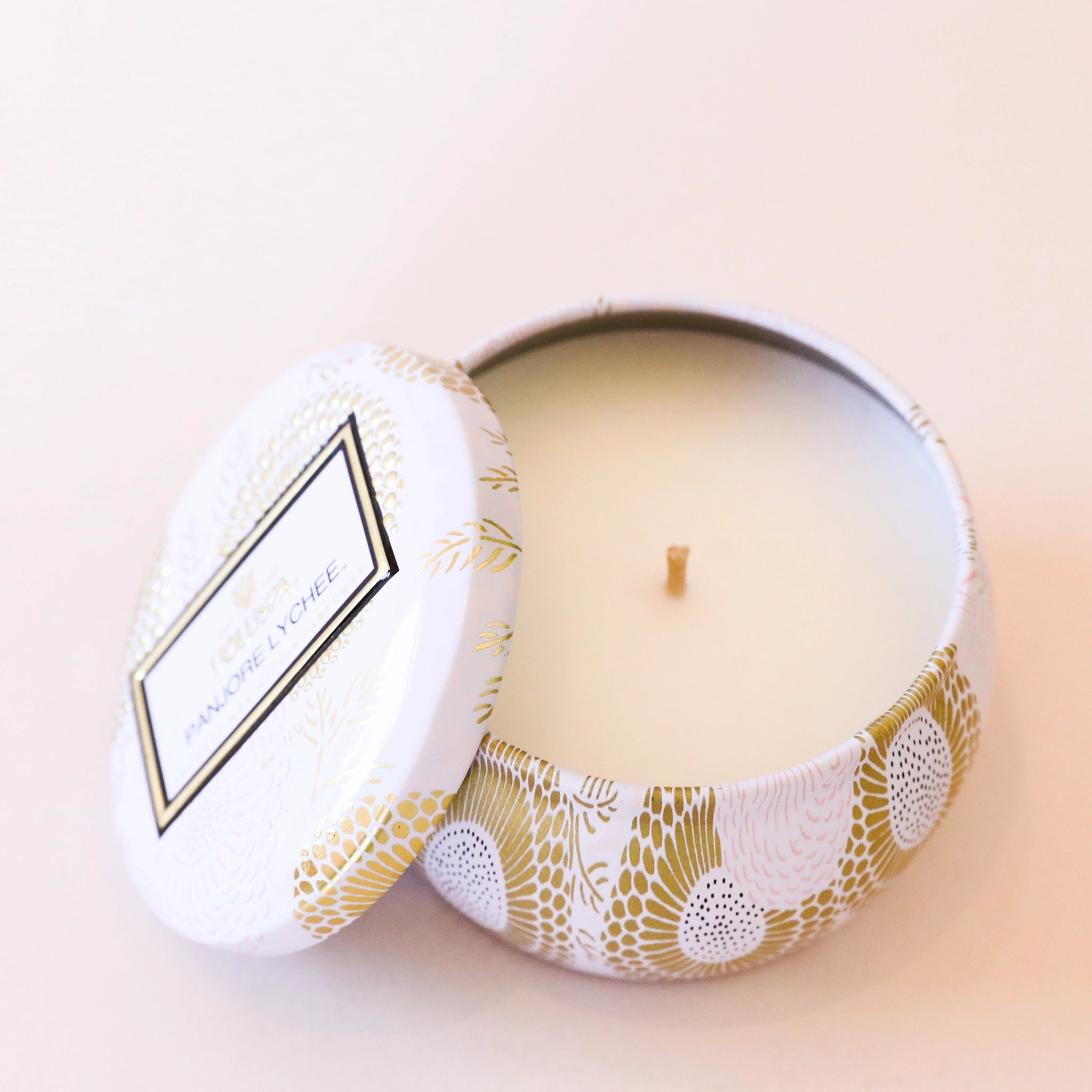 Birds eye view of a short, round tin. Inside the tin is a white candle with a white wick in the center. The tin is light pink with a gold floral pattern. There is a matching lid leaning against the left side of the tin.