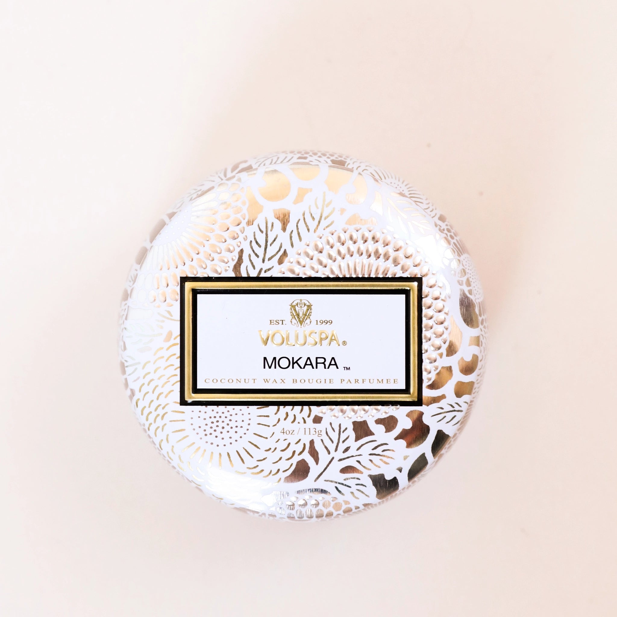 On a cream background is a decorative tin single wick candle with a lid and a label that reads, "Voluspa Mokara"