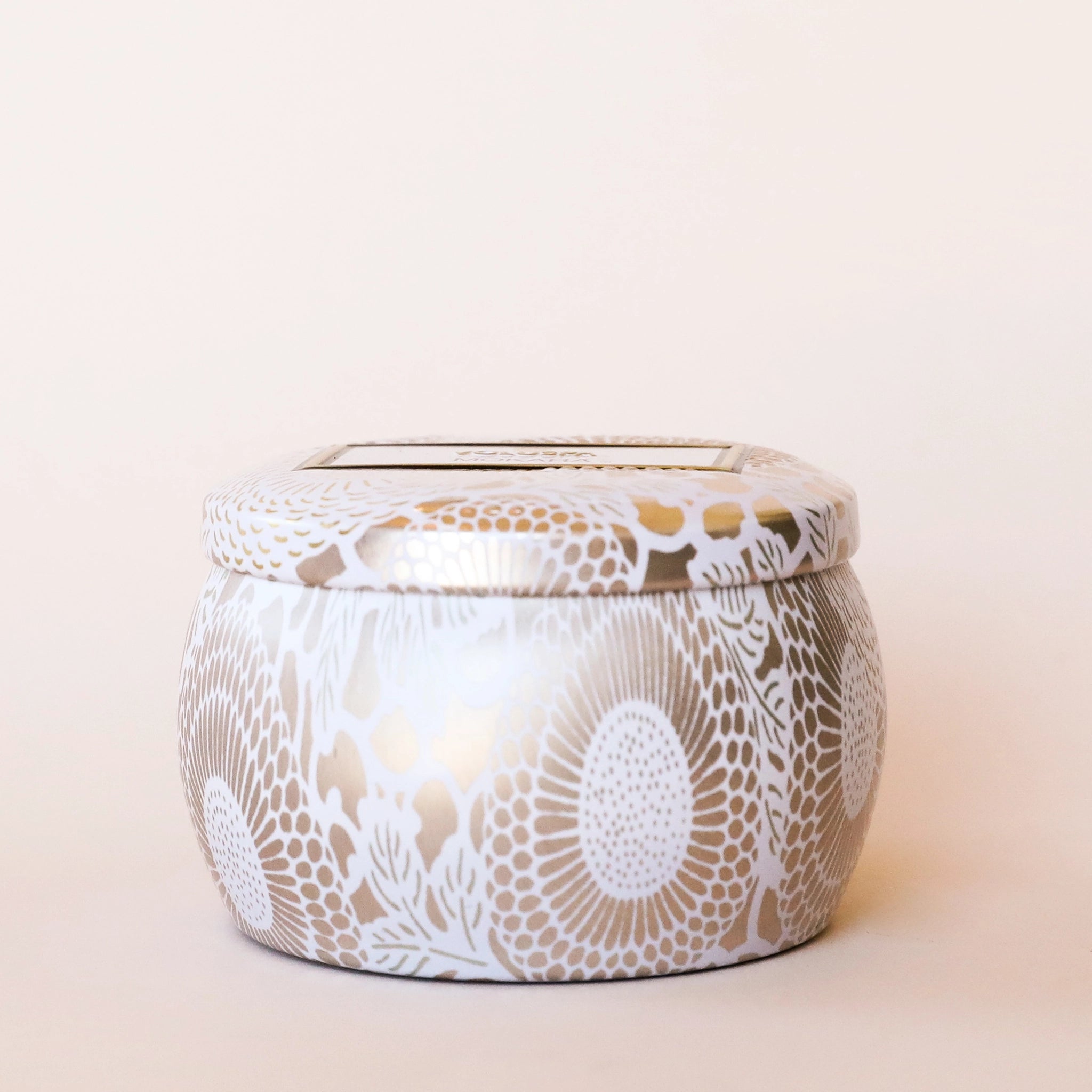 On a cream background is a decorative tin single wick candle with a lid and a label that reads, &quot;Voluspa Mokara&quot;