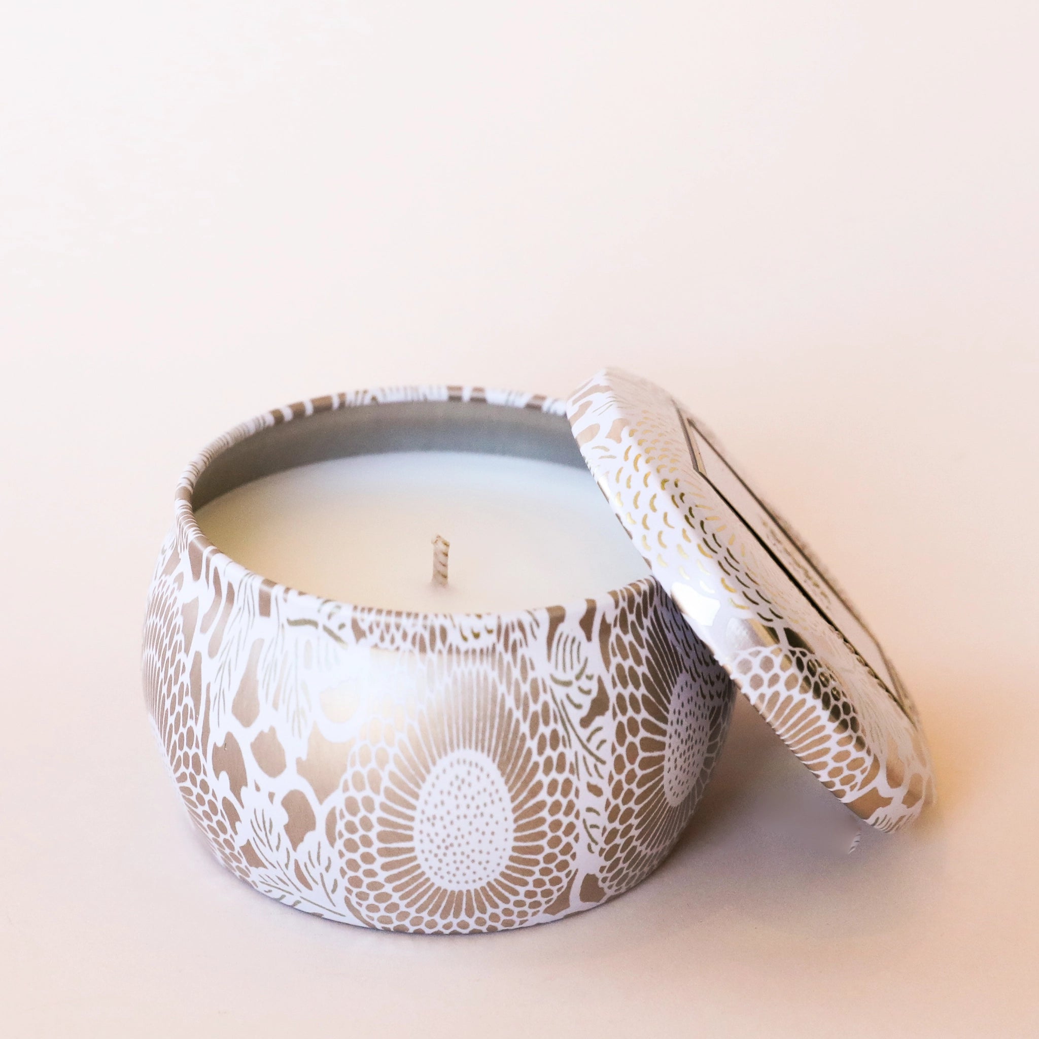 On a cream background is a decorative tin single wick candle with a lid and a label that reads, "Voluspa Mokara"