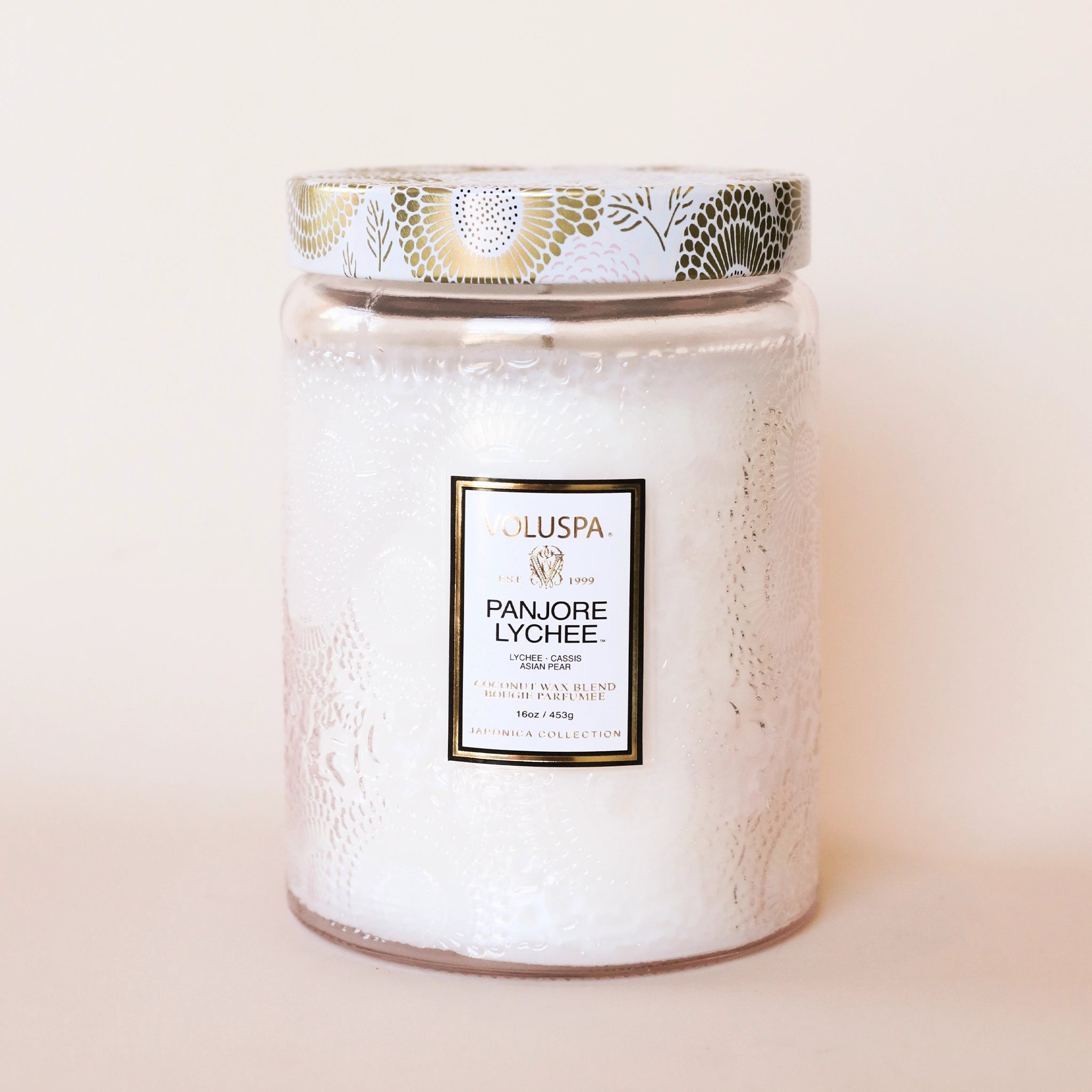 In front of a white background is a tall, light pink glass jar with a short tapered top. The jar has an etched in floral pattern around it. In the middle is a white sticker with a black and gold border. At the top is gold text that reads ‘Voluspa’ and black text that reads ‘Panjore Lychee.’ On top of the jar is a round tin lid. The lid is light pink with a gold floral pattern.
