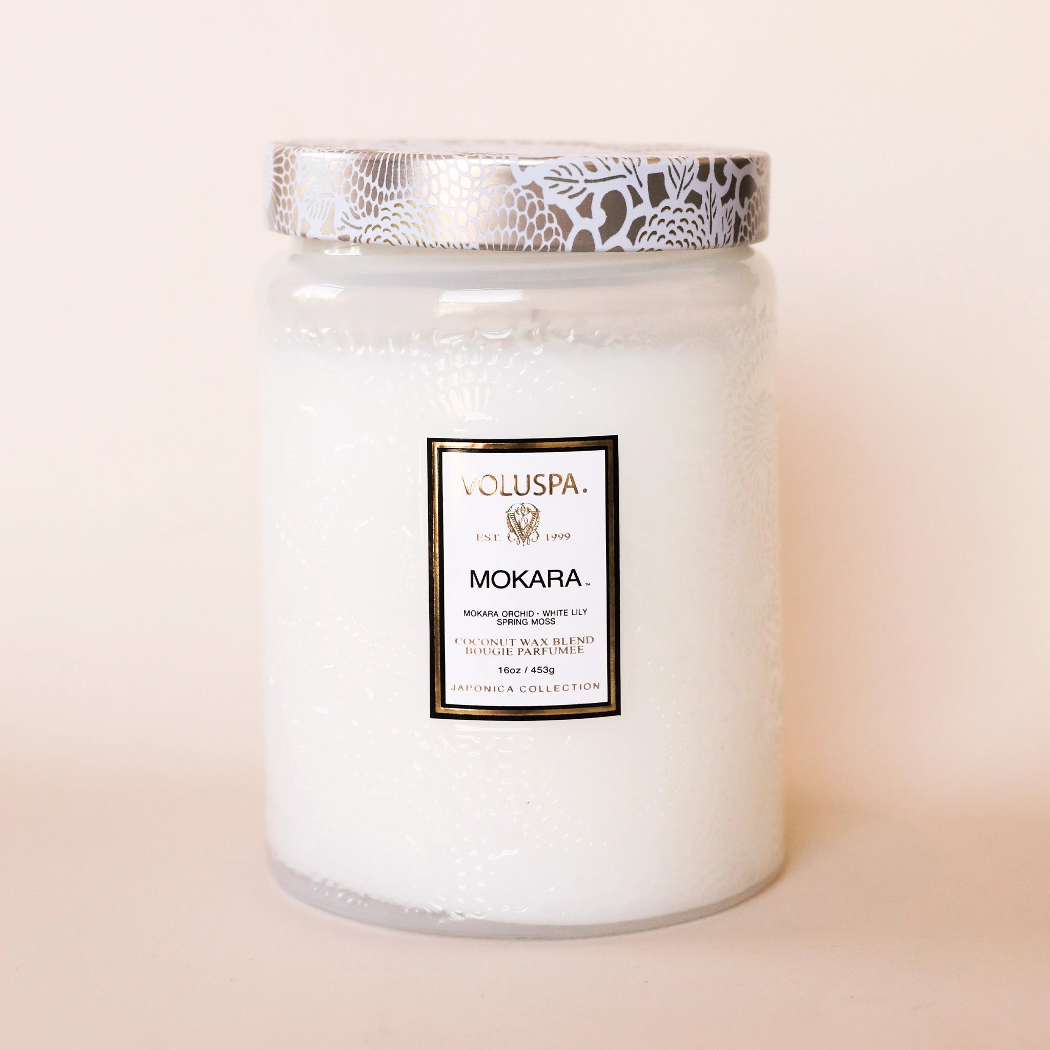 On a white background is a white decorative glass jar with a coordinating lid and a label on the front that reads, "Voluspa Mokara"