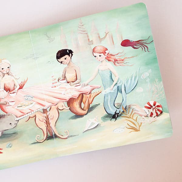 The book open to a page with an illustration of mermaids sitting at a shell shaped table. 