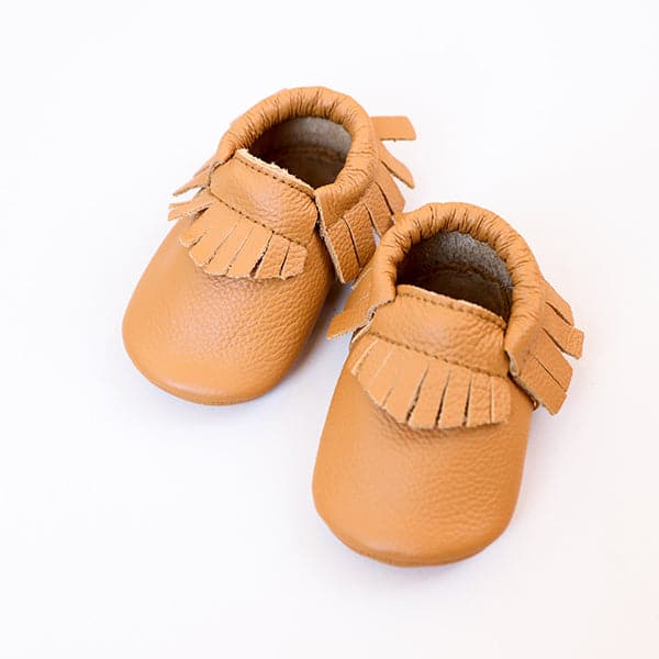 On a white background is a pair of tan baby moccasins that have a fringe detail around the edges.