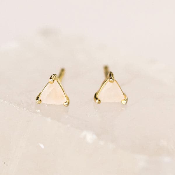 In front of a pink background is a pair of rose quartz studs. The crystal is shaped like a triangle with a gold post on each corner. 