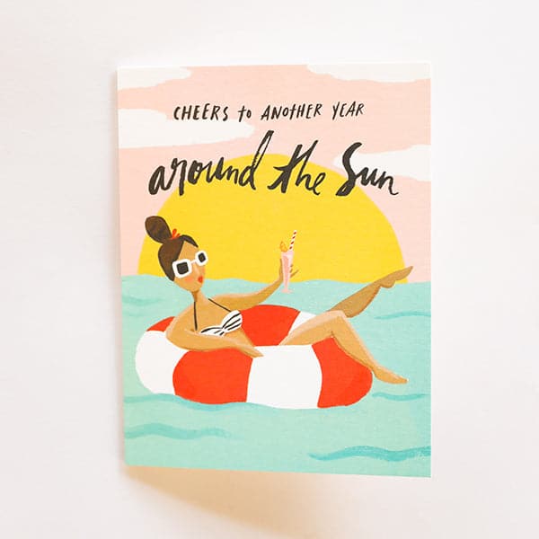 In front of a white background is a card. The card is a drawing of a blue ocean with a pink sky and yellow setting sun. In the pink sky is black text that reads ‘cheers to another year around the sun.’ In the water is a red and white striped inner tube with a girl laying inside. She is wearing white sunglasses and holding a pink drink. 