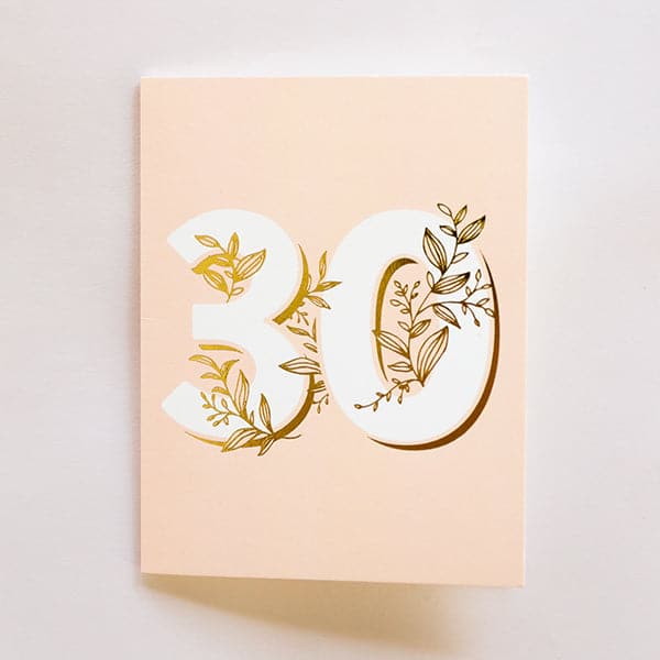 A light pink card with a cream colored envelope and big white numbers that read, &quot;30&quot; along with gold foiled dainty vines that wrap through and around the numbers.