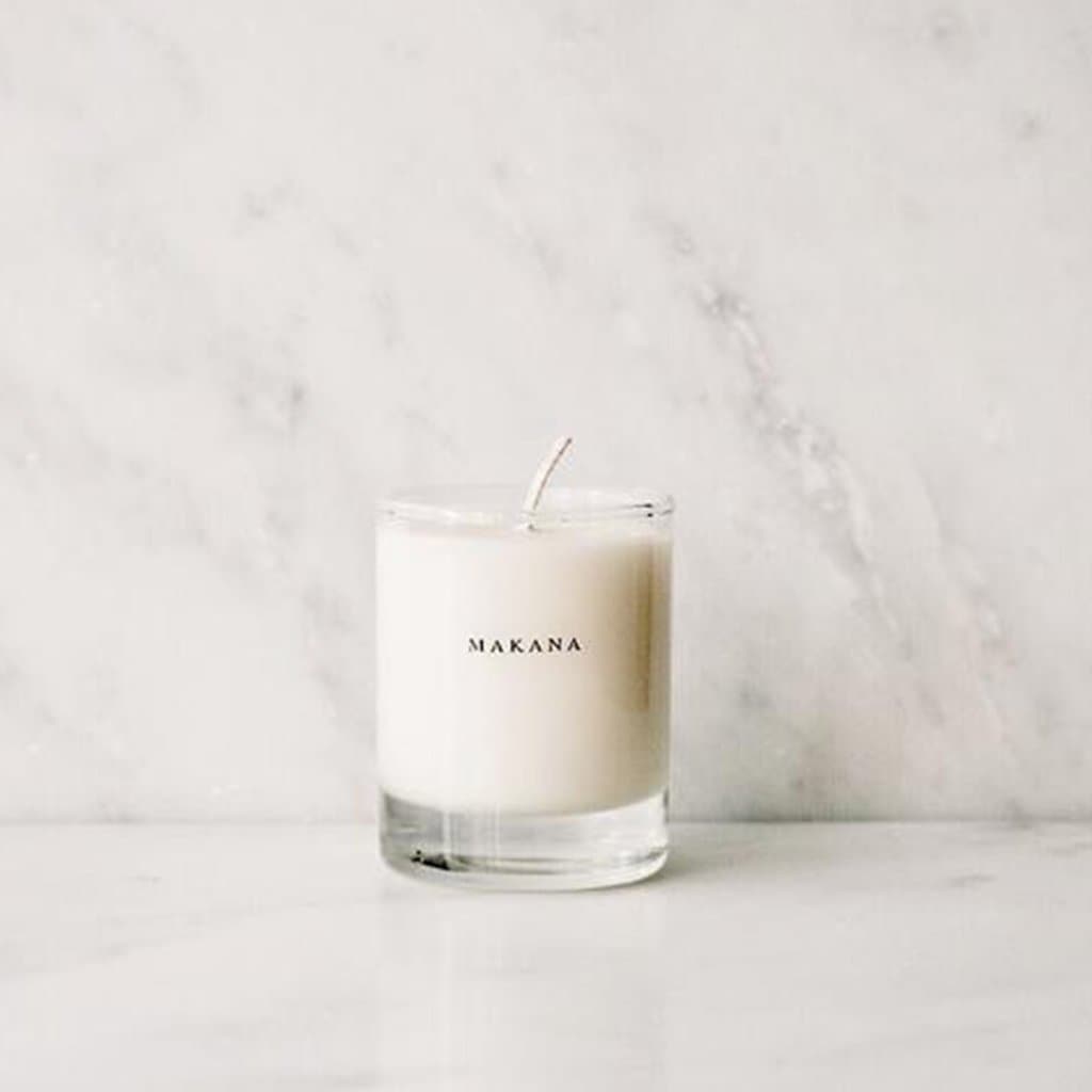 In front of a white and gray marbled wall is a round glass jar. Inside the jar is a white candle with a white wick in the center. On the front is black text that reads ‘Makana.’