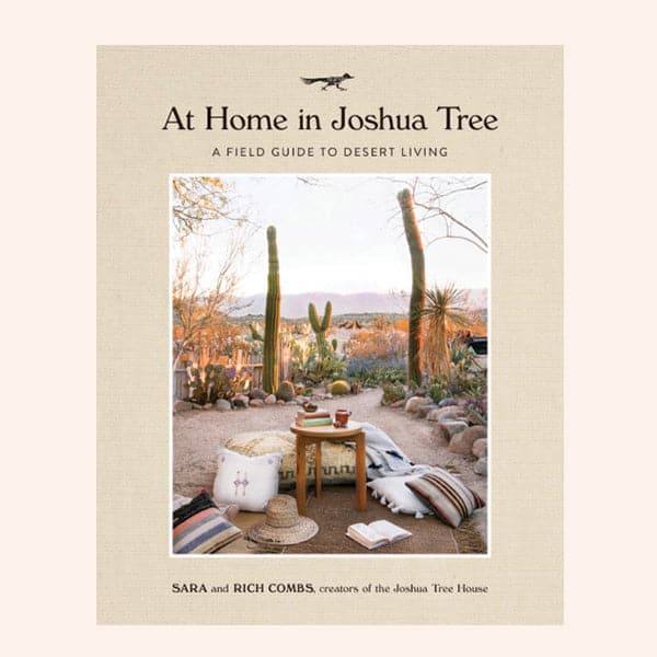 A natural colored book titled "At Home in Joshua Tree - a field guide to desert living - Sara and Rich Combs creators of the Joshua Tree House" with image of a desert scene with small wooden table with books and mugs, and pillows laying on the ground.